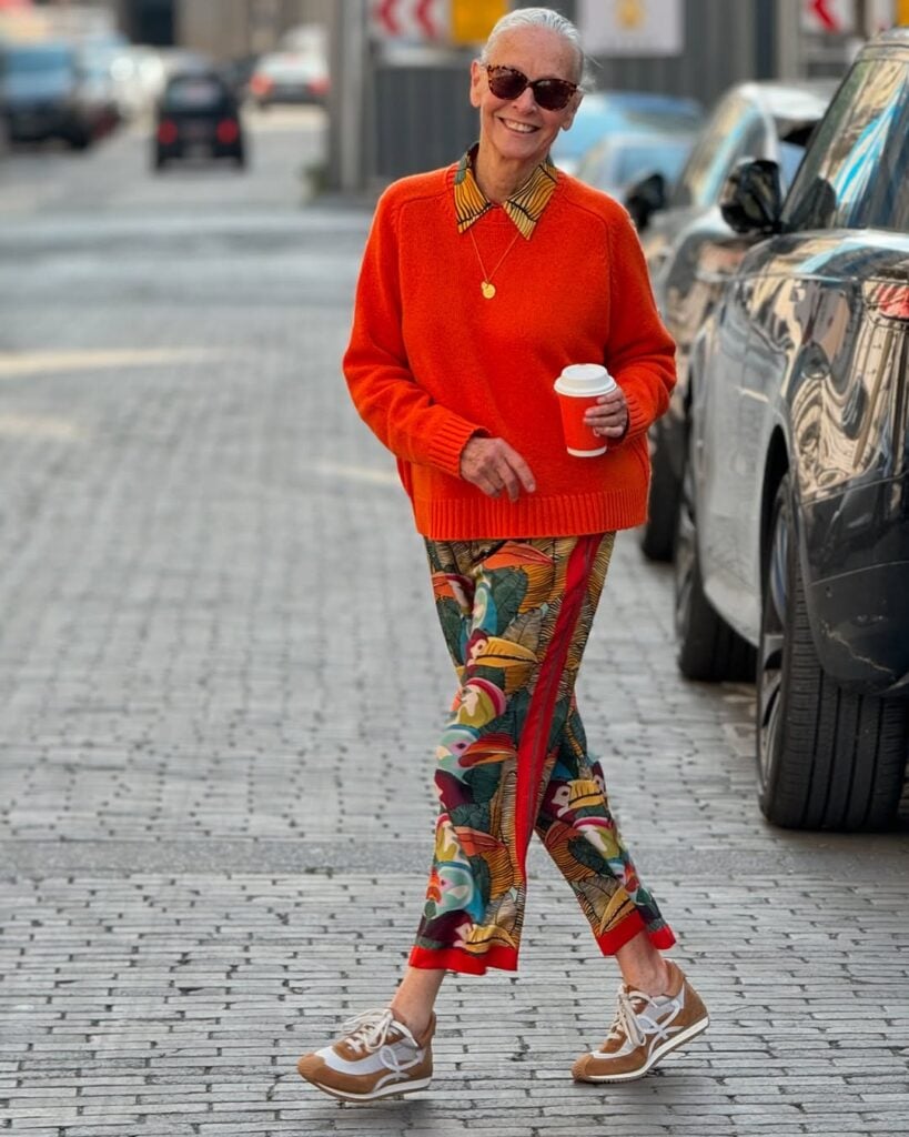 Mulher com mais de 50 anos. Ela usa calça com estampa, suéter laranja tênis.