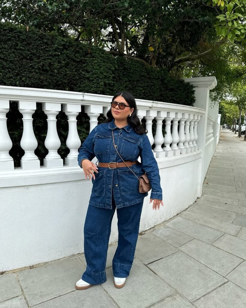 Mulher jovem, de cabelo escuro e pele clara. Ela usa look all jeans, tênis, cinto marrom e óculos de sol.