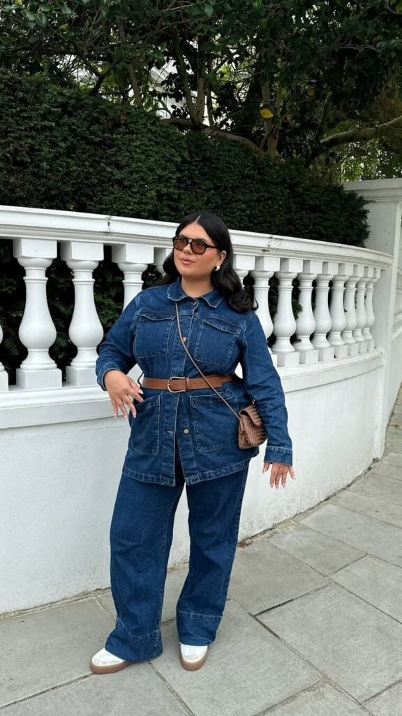 Mulher jovem, de cabelo escuro e pele clara. Ela usa look all jeans, tênis, cinto marrom e óculos de sol.