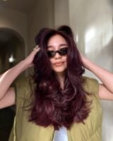 Mulher de pele clara, com blusa branca e colete verde, acessórios em dourado, óculos de sol e cabelo burgundy