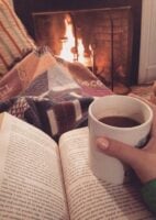Pessoa sentada com manta e livro sobre o colo, segurando uma caneca de chocolate quente