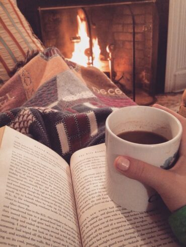 Pessoa sentada com manta e livro sobre o colo, segurando uma caneca de chocolate quente