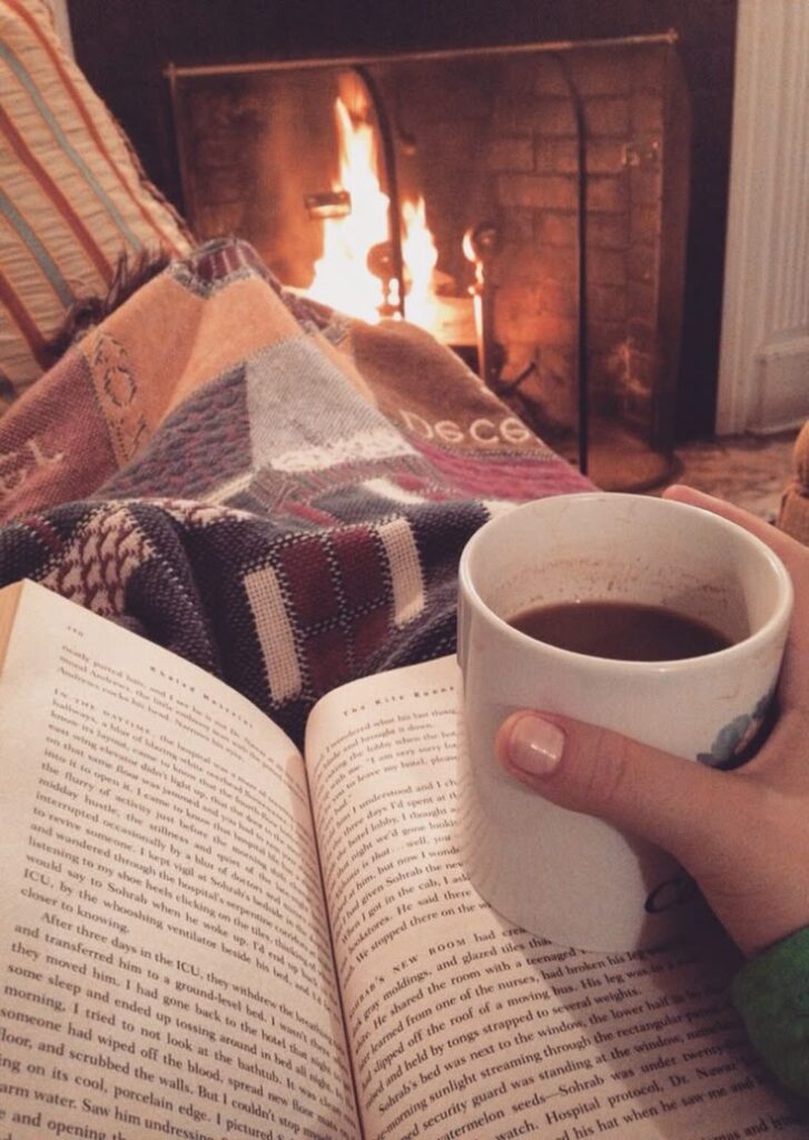 Pessoa sentada com manta e livro sobre o colo, segurando uma caneca de chocolate quente