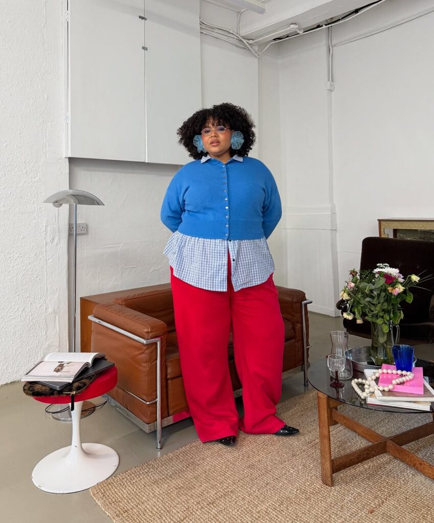 Mulher jovem com suéter azul, camiza xadrez azul e branca, calça vermelha e sapato preto.