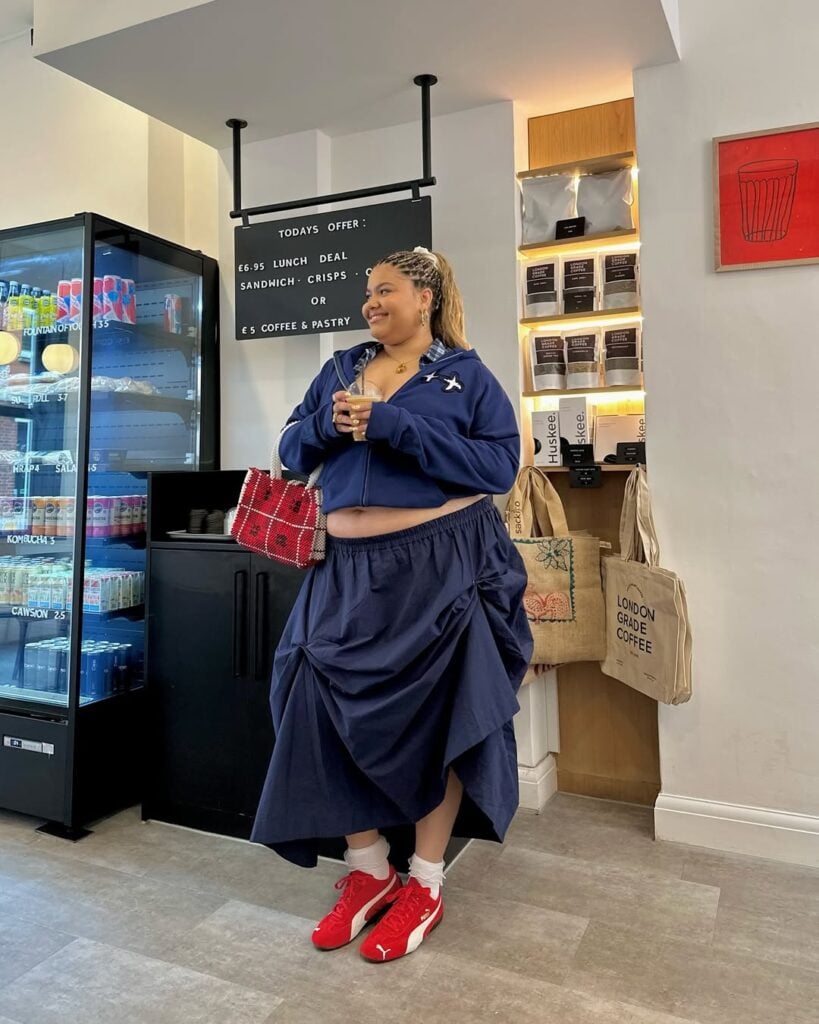 Mulher jovem com look azul marinho composto por casaco e saia longa. Ela está segurando uma bolsa vermelha e usa tênis Puma Speedcat vermelho.