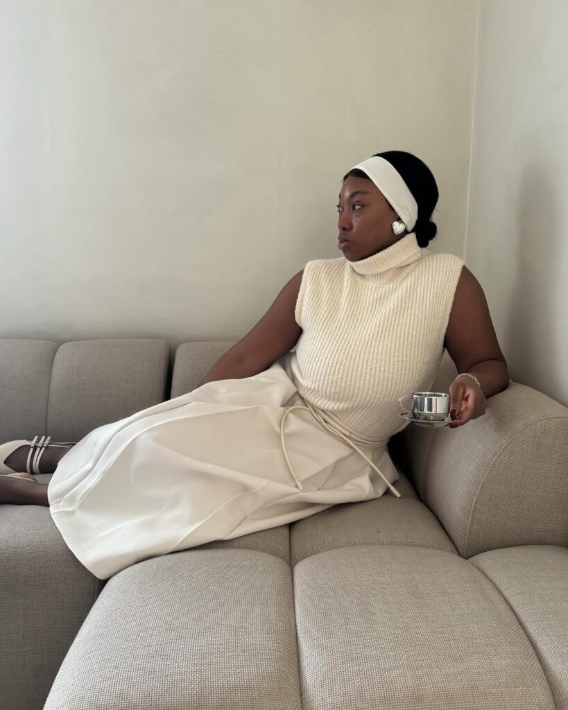 Mulher de pele preta, sentada em um sofá, com look off white e faixa de cabelo na mesma cor
