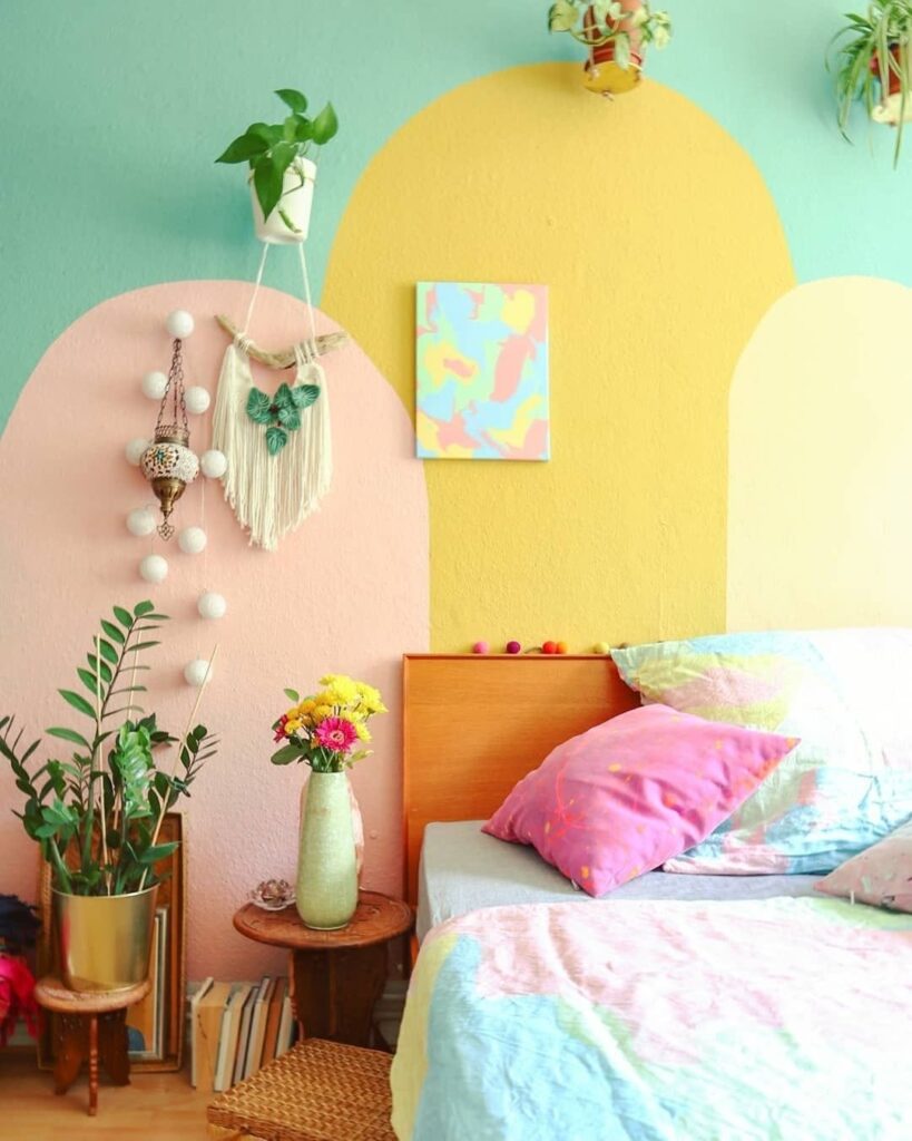 Quarto com cama e mesa de cabeceira em madeira e pintura colorida em rosa, amarelos e verde atrás da cama