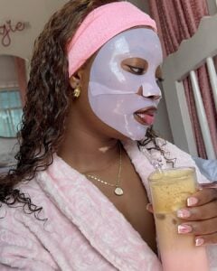 Mulher jovem de pele negra e cabelos cacheados. Ela usa faixa de cabelo, máscara facial e roupão.