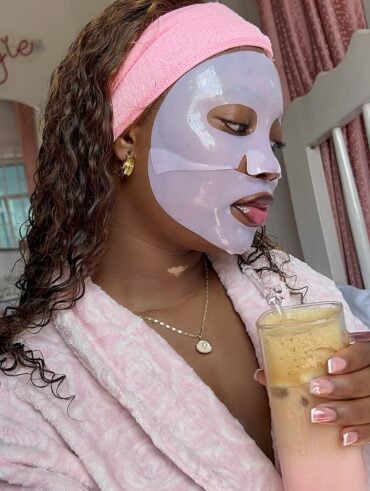Mulher jovem de pele negra e cabelos cacheados. Ela usa faixa de cabelo, máscara facial e roupão.