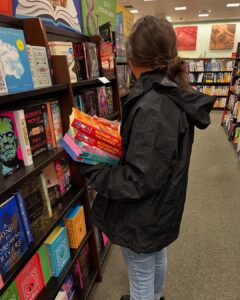 Mulher de pele clara, com cabelo castanho, casaco preto e calça jeans. Ela está segurando uma pilha de livros