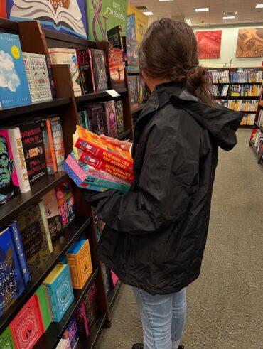 Mulher de pele clara, com cabelo castanho, casaco preto e calça jeans. Ela está segurando uma pilha de livros