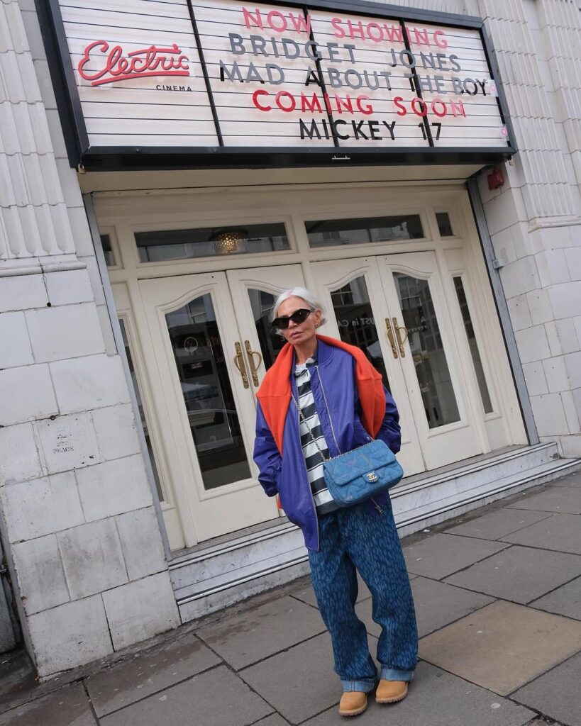 Mulher com mais de 50 anos, cabelos curtos e pele clara. Ela está parada na frente de um letreiro de cinema e usa roupas coloridas.