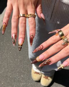 Mãos de pele clara, com anéis em dourado e unha amendoada com multi decorações e charms em dourado