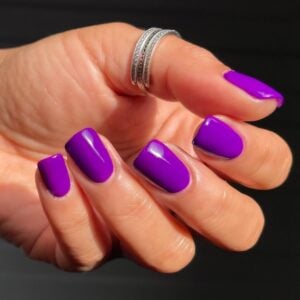 Mão de pele clara, com anel em prata e unha quadrada com esmalte roxo neon