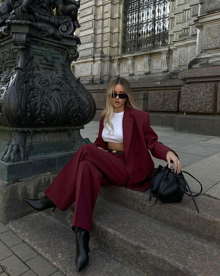 Mulher adulta com cabelos loiros e pele clara. Ela está sentada em degraus na rua e usa conjunto de calça e blazer na cor vermelho-cereja.