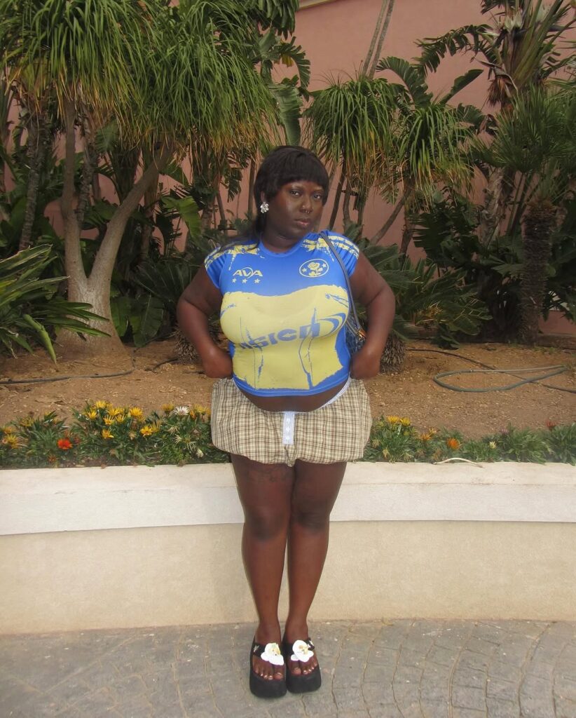 Mulher jovem, plus size e de pele negra. Ela usa camiseta, saia balonê e tamanco.