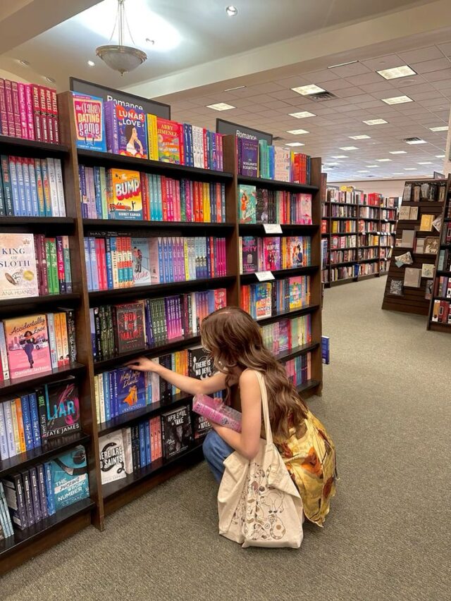 Moça jovem com cabelos longos na frente de uma livraria com uma estante cheia de livros.