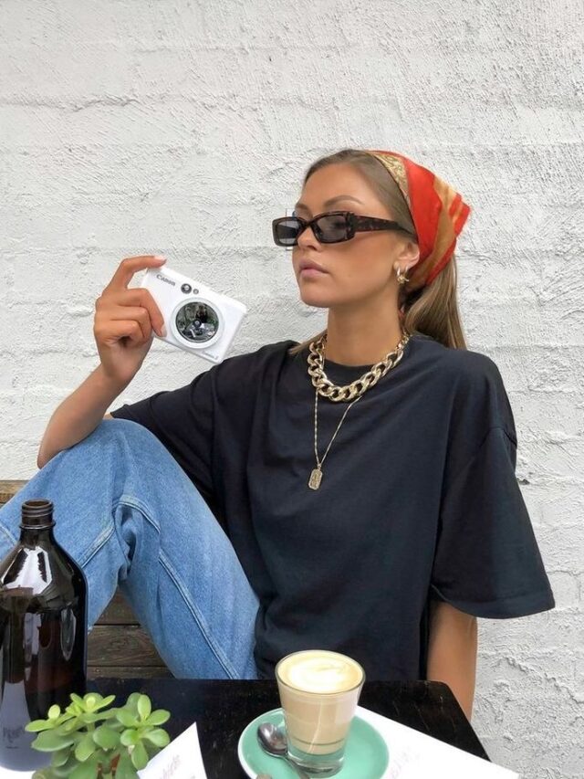 Mulher jovem branca com lenço no cabelo liso. Ela usa óculos de sol, acessórios dourados, camiseta preta básica e calça jeans