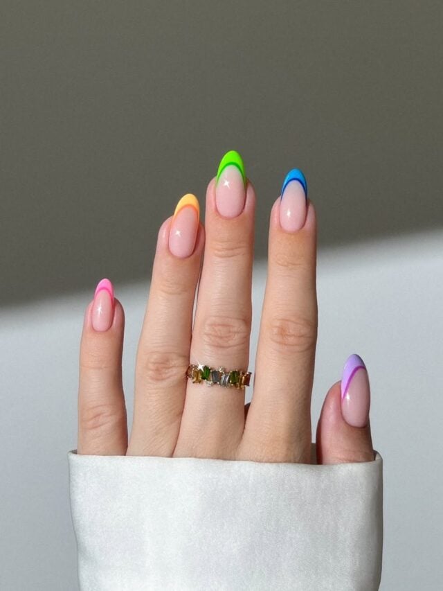 Mão branca com unhas longas e um anel de pedras. A esmaltação é uma francesinha colorida nas cores rosa, amarelo, verde, azul e lilás, uma cor em cada unha.