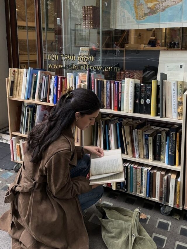 Mulher jovem e magra de pele clara e com cabelos longos e escuros. Ela está na frente de uma estante cheia de livros e folheia um deles.