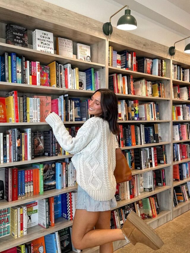 Mulher jovem de pele clara com suéter branco e vestido listrado. Ela está na frente de uma estante de livros.