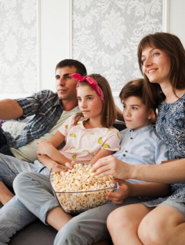 Família comendo pipoca e assistindo à televisão sentada em um sofá.
