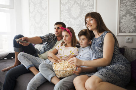Família comendo pipoca e assistindo à televisão sentada em um sofá.
