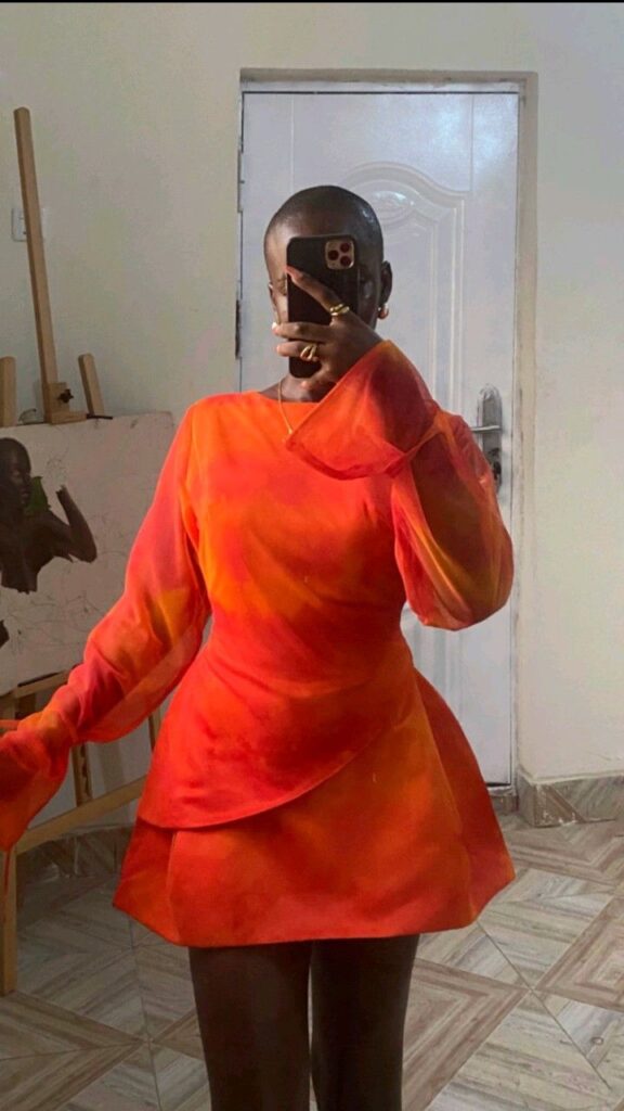 Mulher negra na frente do espelho com o celular. Ela veste um vestido de mangas longas laranja e vermelho.