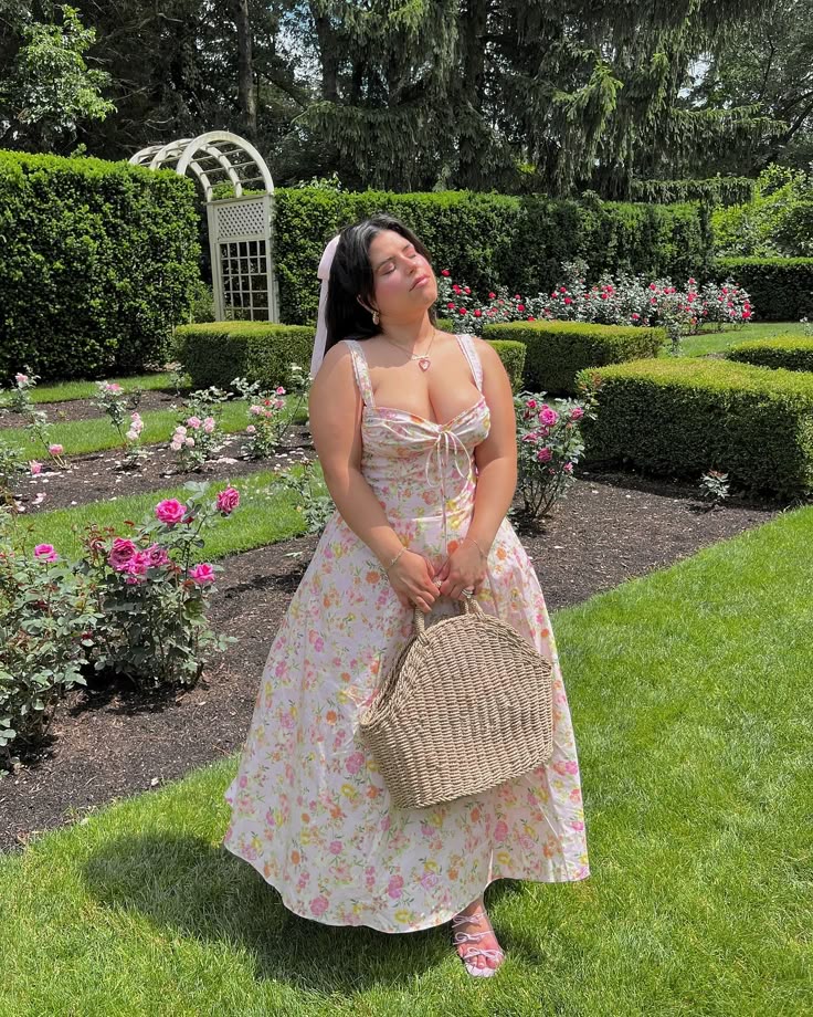 Mulher jovem com pele clara em um jardim florido. Ela usa vestido com estampa floral e está segurando uma bolsa de palha.