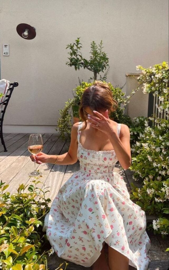 Mulher jovem, magra e de pele clara. Ela está sentada com a perna cruzada e usa look floral.