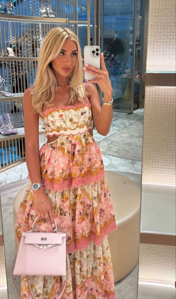 Mulher jovem e loira. Ela veste um vestido floral longo em tom rosa.