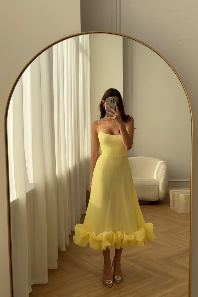 Mulher jovem e magra na frente do espelho tirando foto. Ela veste um vestido midi amarelo e usa sandália com bico fino.