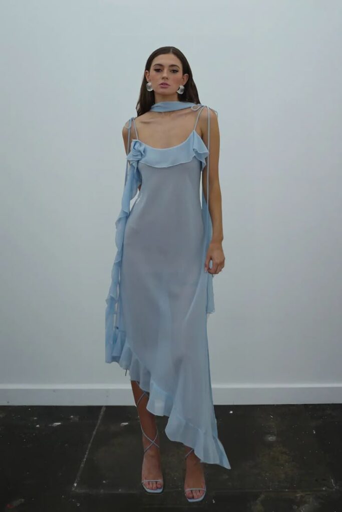 Mulher adulta com pele clara e cabelos escuros. Ela usa vestido azul claro midi e sandálias azuis.