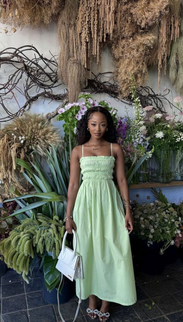 Mulher jovem e magra de pele escura. Ela usa vestido verde liso e segura uma bolsa branca nas mãos.