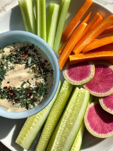 Verduras e frutas dispostas em um prato branco com um molho em uma cumbuca