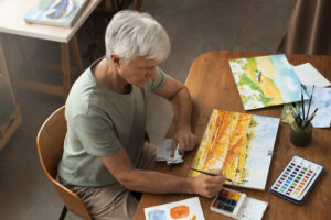 Homem com cabelos grisalhos sentado pintando uma paisagem sobre um papel