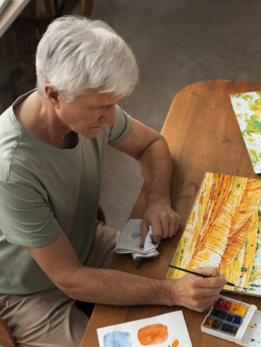 Homem com cabelos grisalhos sentado pintando uma paisagem sobre um papel