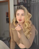 Mulher de pele clara, com top de manda longa Mocha Mousse, cabelo loiro em ondas. Ela está passando gloss
