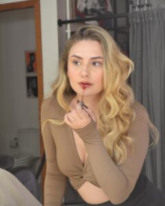 Mulher de pele clara, com top de manda longa Mocha Mousse, cabelo loiro em ondas. Ela está passando gloss