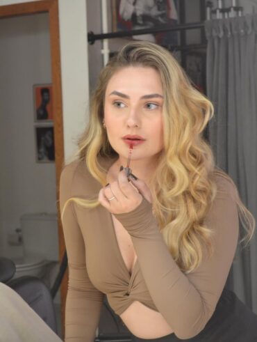 Mulher de pele clara, com top de manda longa Mocha Mousse, cabelo loiro em ondas. Ela está passando gloss