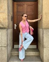 Mulher jovem, magra e de pele clara. Ela usa colete rosa, calça jeans clara e salto alto rosa.