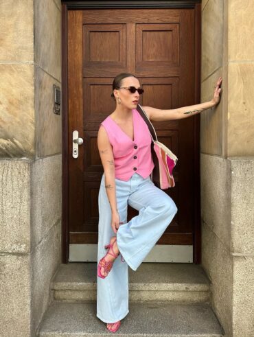 Mulher jovem, magra e de pele clara. Ela usa colete rosa, calça jeans clara e salto alto rosa.