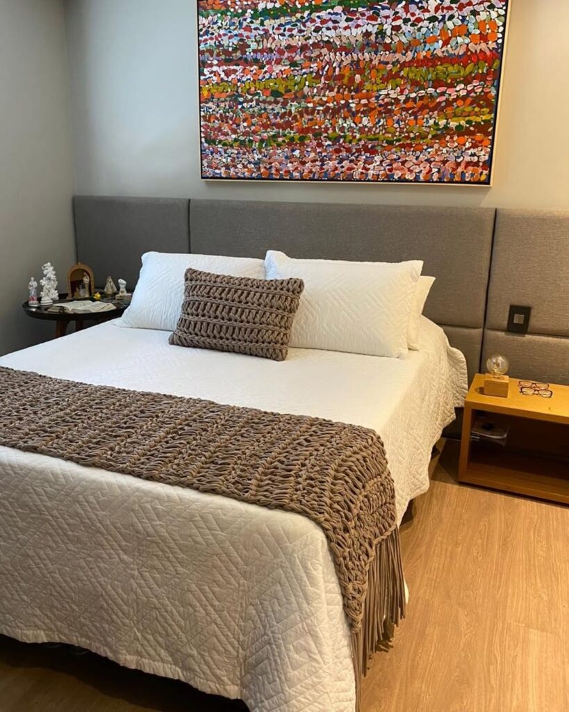 Quarto com cabeceira estofada cinza, roupa de cama branca e manta e almofada de crochê