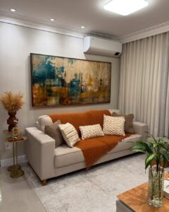 Sala de estar com sofá claro, manta terrosa e quadro com pintura abstrata na parede