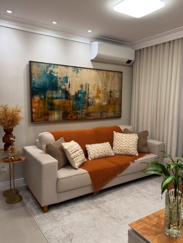 Sala de estar com sofá claro, manta terrosa e quadro com pintura abstrata na parede