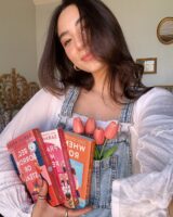 Mulher de pele clara, com cabelo castanho, blusa branca e jardineira jeans. Ela está segurando livros