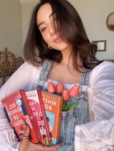Mulher de pele clara, com cabelo castanho, blusa branca e jardineira jeans. Ela está segurando livros