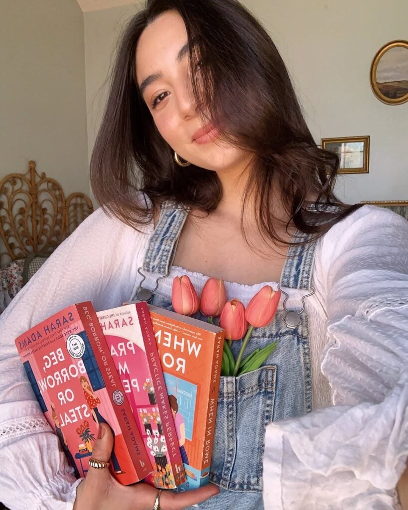 Mulher de pele clara, com cabelo castanho, blusa branca e jardineira jeans. Ela está segurando livros