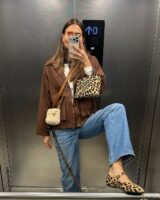 Mulher jovem e magra com look marrom e calça jeans. Ela usa bolsa e sapatilha animal print.