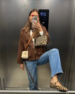 Mulher jovem e magra com look marrom e calça jeans. Ela usa bolsa e sapatilha animal print.
